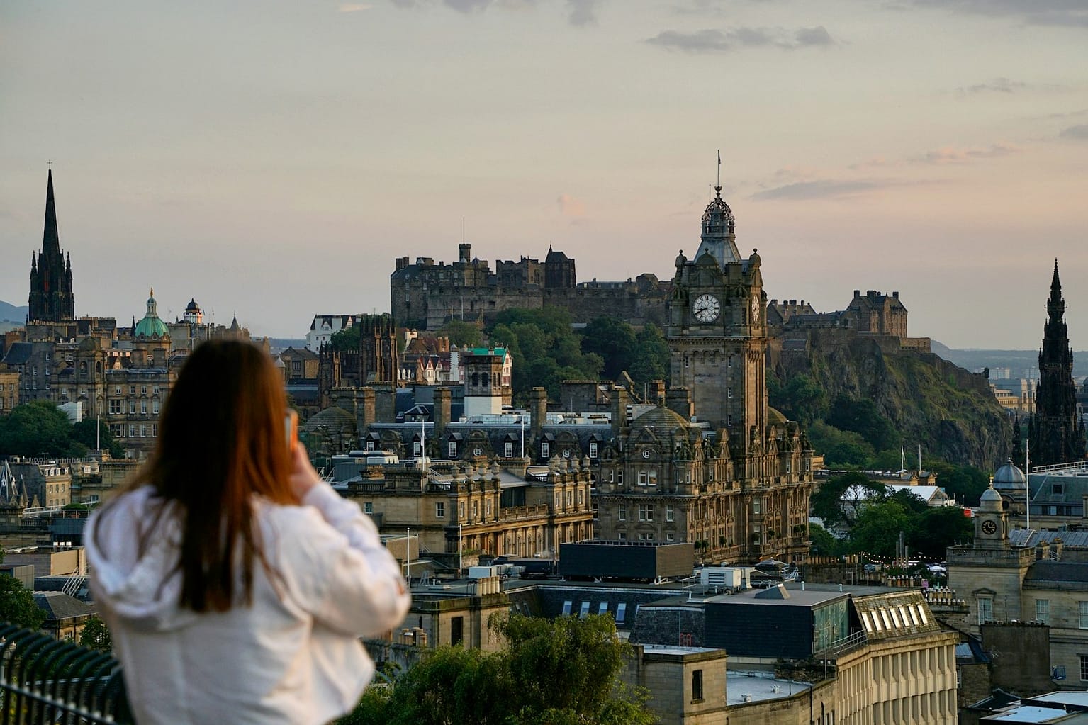 woman in white shirt looking at the city of Edinburgh and taking a photo during daytime